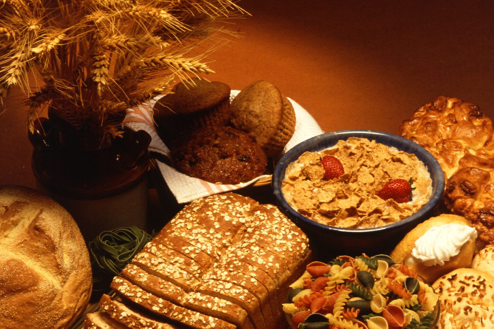 Bread, a sliced wholegrain loaf, muffins, a bowl of bran flakes, pasta and other baked goods on a table beside a sheaf of ripe wheat.