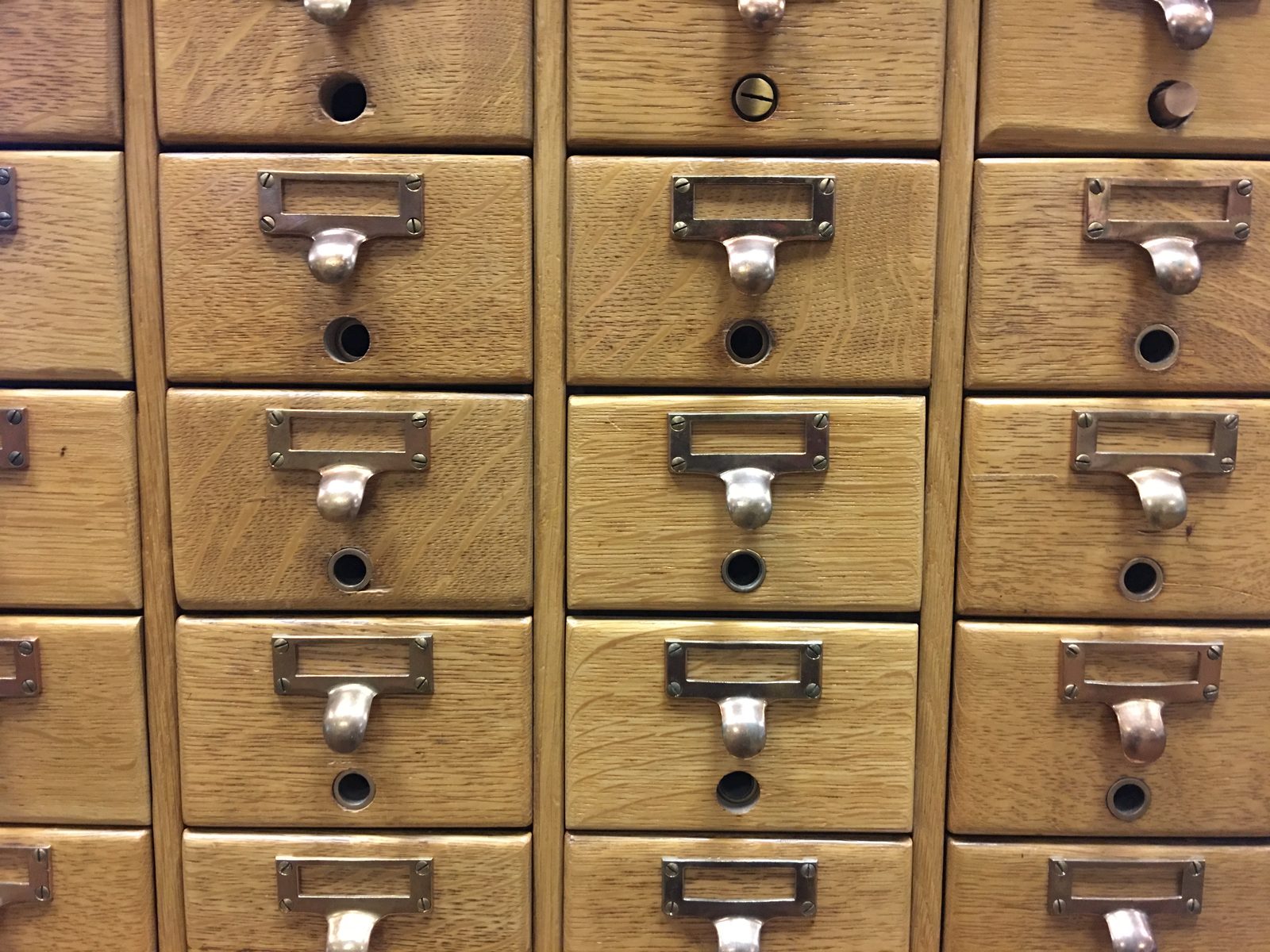 A wooden library card catalogue: rows of small oak drawers, each with a brass label holder above a pull handle.