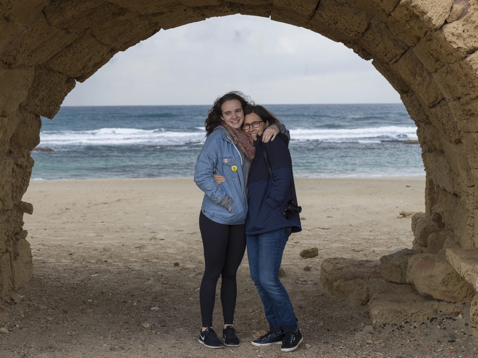 A young woman and an older woman in glasses stand arm in arm under a stone arch on a beach, smiling at the camera, with the sea behind them.