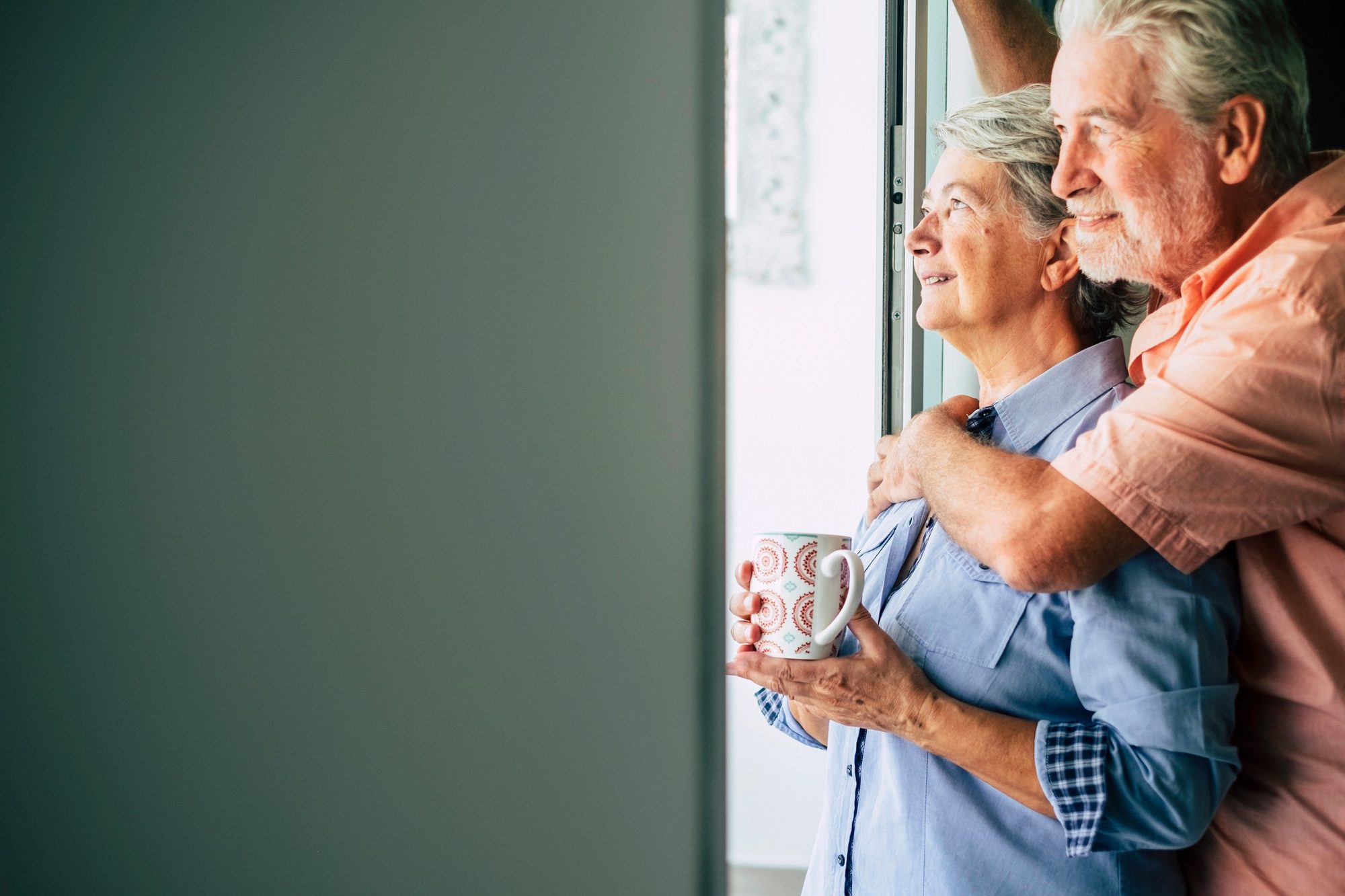 An older couple look out of a window, smiling. He holds her from behind, and she holds a mug in both hands.