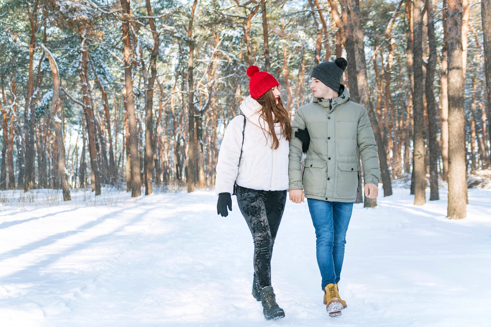 A young couple in winter hats walk arm in arm through a snowy pine forest, looking at each other.