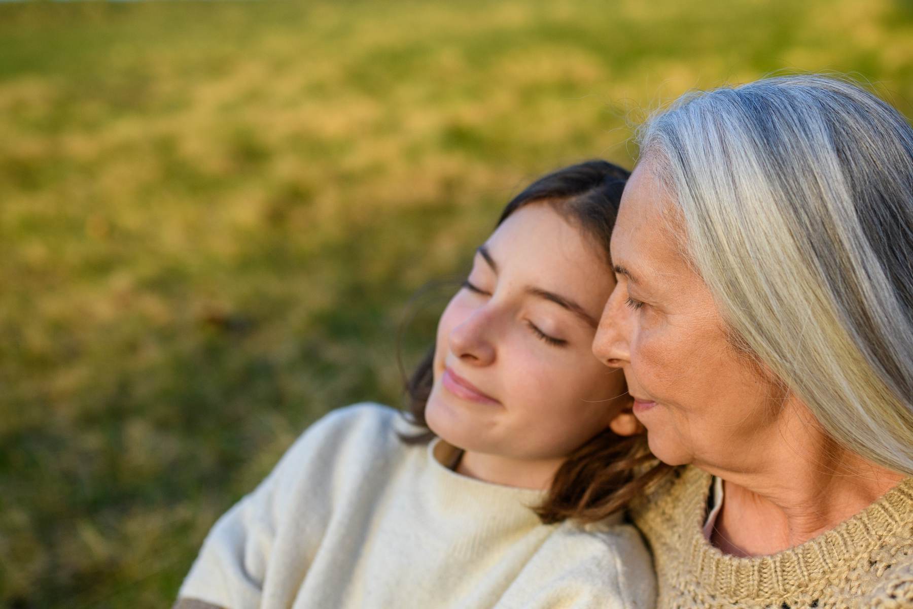 A teenage girl and an older woman with long grey hair rest cheek to cheek outdoors. Both have their eyes closed and are smiling.