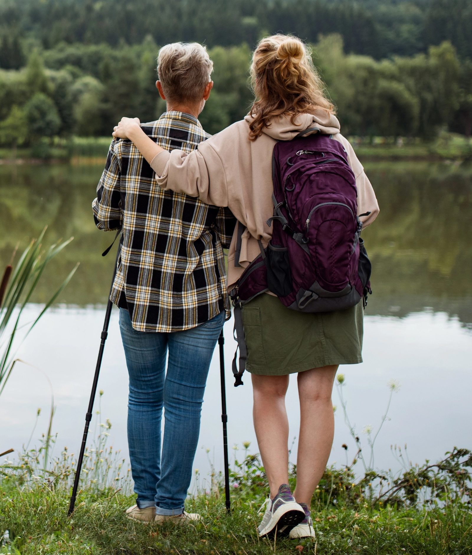 Seen from behind, a younger woman with a backpack has her arm around an older woman holding trekking poles. They stand at the edge of a lake, looking across to wooded hills.