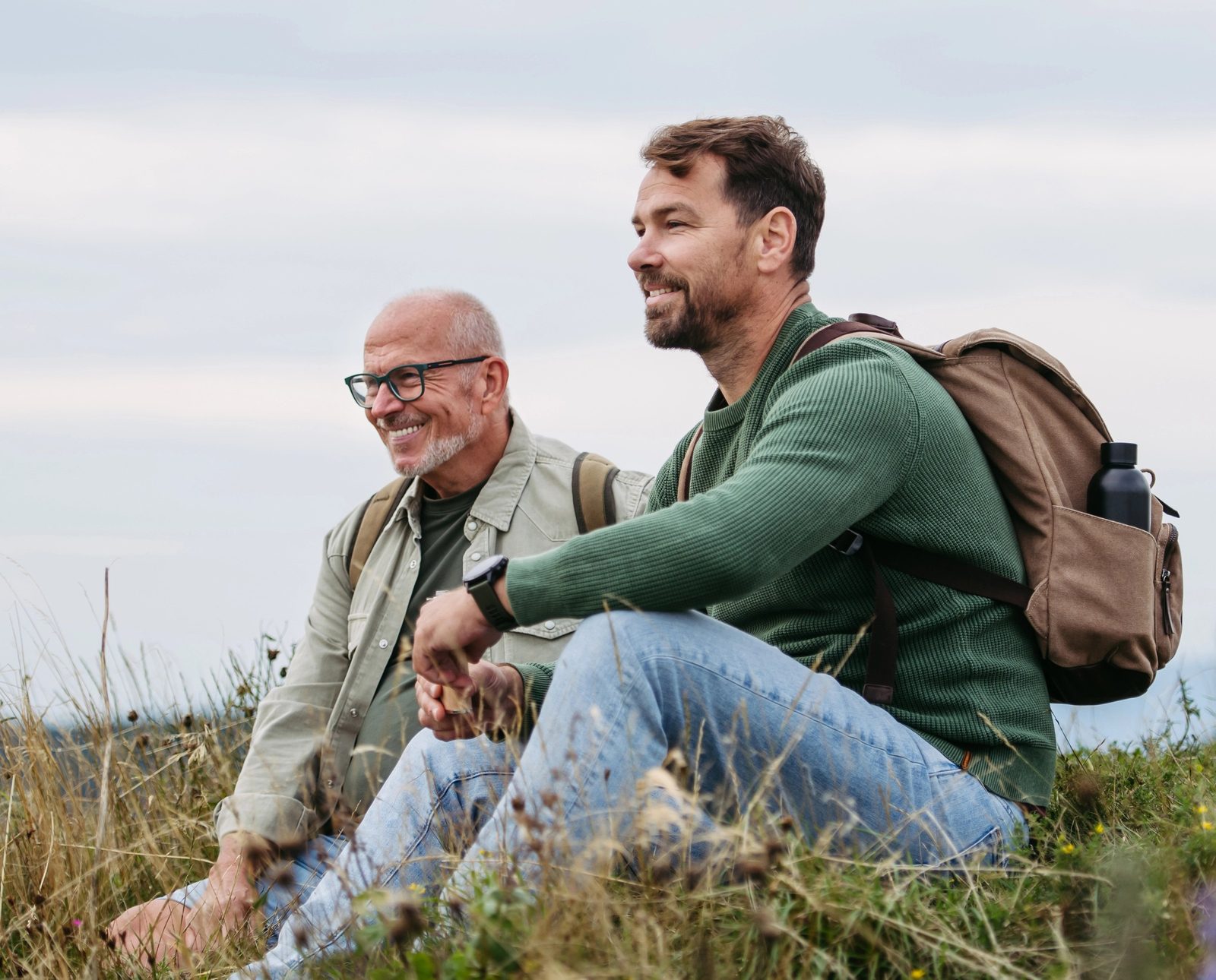 An older man in glasses and a younger man with a backpack sit side by side in tall grass on a hilltop, smiling as they look into the distance.